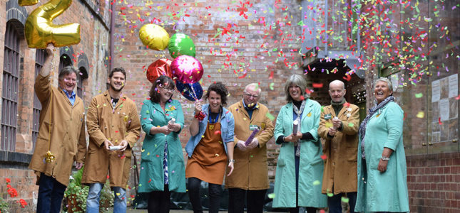 Eight members of the team in Newman Brothers uniforms, some hold balloons, one of which is shaped like the number 2. They fire confetti canons and laugh.