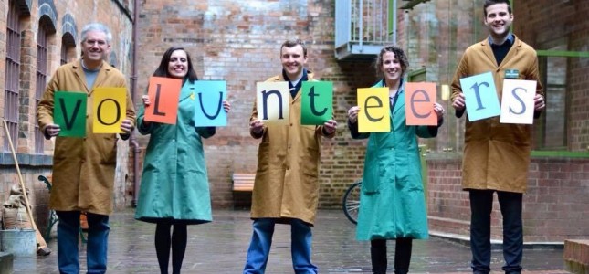 Volunteers stand in a row, holding up coloured sheets of paper spelling out the word "volunteers".