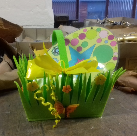 Green Easter basket with a large spotted egg sits on bench surrounded by packages and coffin handles