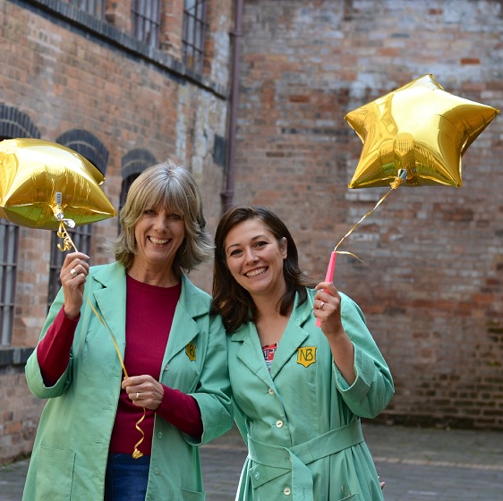 Two happy volunteers in green Newman Brothers coats holding star shaped balloons