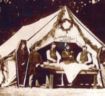 Civil war embalming taking place outside a white tent. There are simple six sided coffins standing on both sides of the tent with soldiers stood inside them.