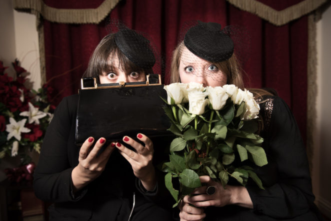 Two women in funeral dress, face the camera with wide open eyes, while hiding the bottom halves of their faces with the bouquet of roses and handbag they are holding.