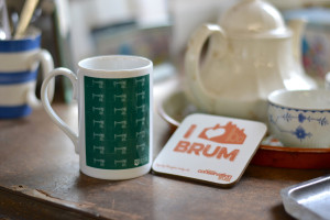 cw shop mug white and green stands next to an i love brum coaster.
