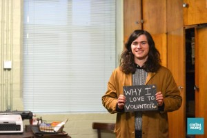 Volunteer in brown overall holds a sign that reads "why i love to volunteer"