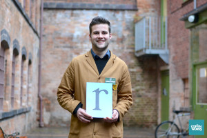 volunteer in brown overall stands in courtyard hold an a4 sized sign showing the letter r in black on a green background