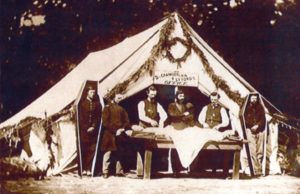 Civil war embalming taking place outside a white tent. There are simple six sided coffins standing on both sides of the tent with soldiers stood inside them.