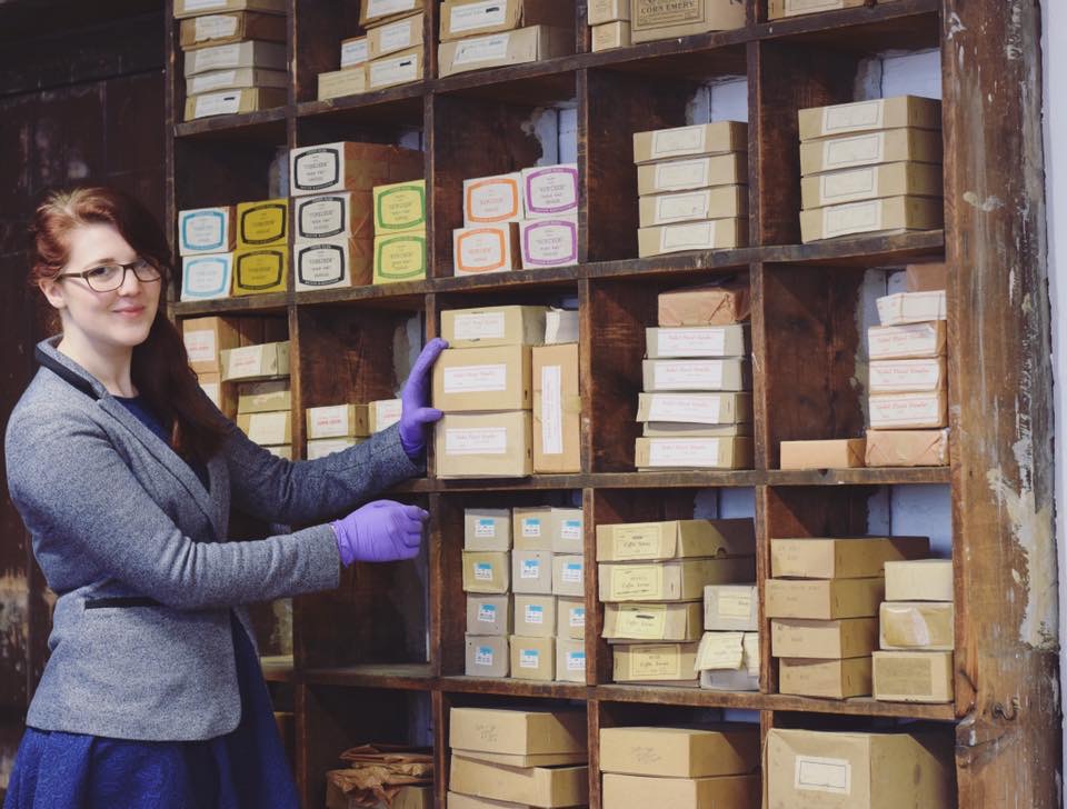 On then left stands a woman with long red hair and grey jacket. Her hand rests on one of the various boxes of coffin handles resting on shelves behind. She wears protective purple gloves.