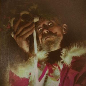 A man with grey moustache and beard wearing Father Christmas clothing looks down at the viewer, he holds a mask in front of his face and is lit from below which gives a spooky feel.