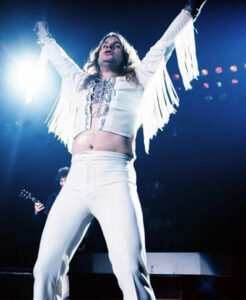 Rock star in tight white clothing white on stage with arms in the air.