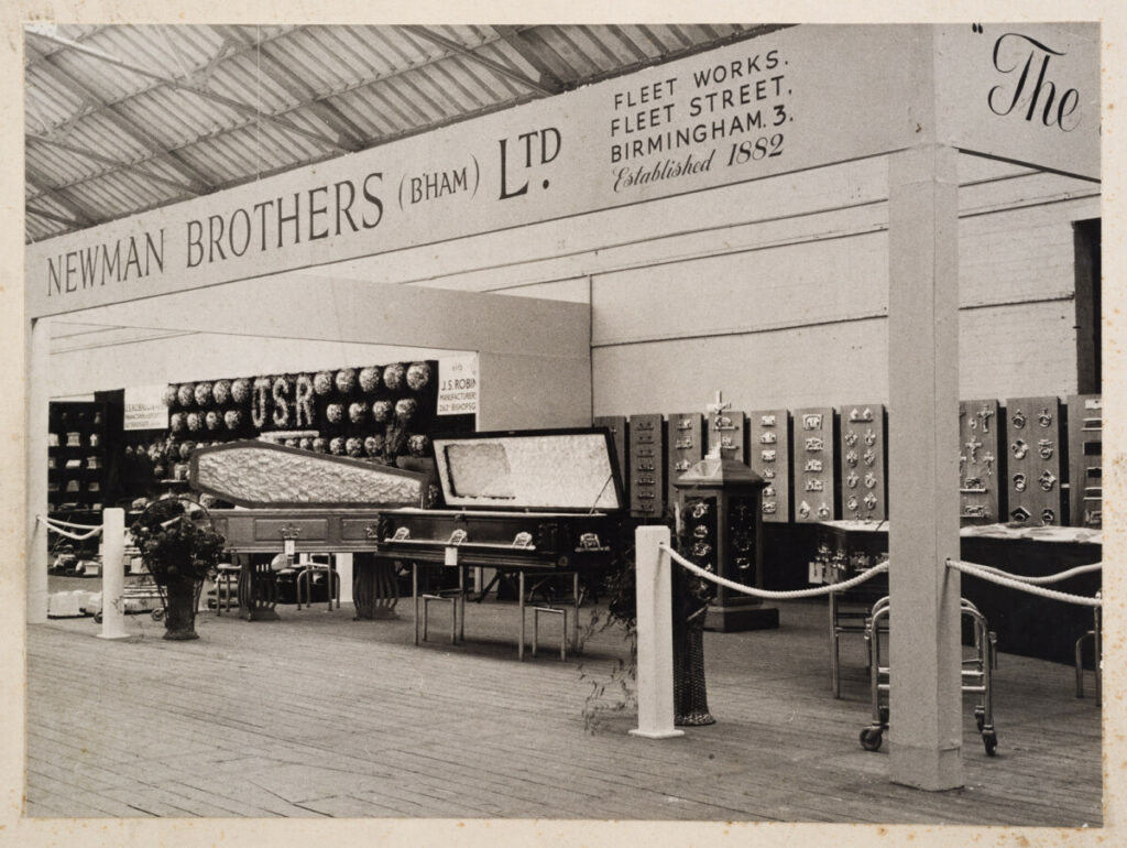 Black and white image of Newman Brothers' Trade show display. We see a coffin a casket and stands of coffin furniture