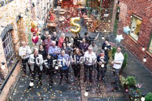 a group of about 20 people stand in coffin works' courtyard. Some holding inflated balloons, confetti falls around them. One balloon is in the shape of a golden number 5, other balloons are silver and are in the shape of letters spelling "coffin works"