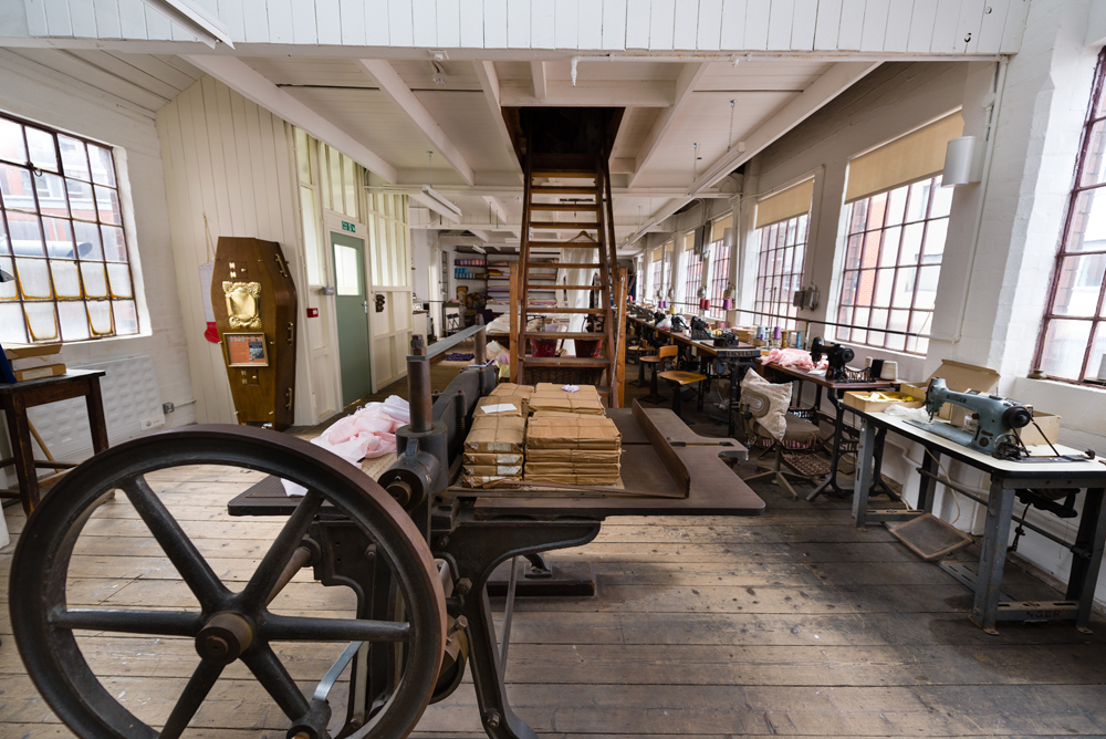 A room featuring windows on the right hand wall, the crimping machine in the foreground. stair descend from the ceiling in the centre of room