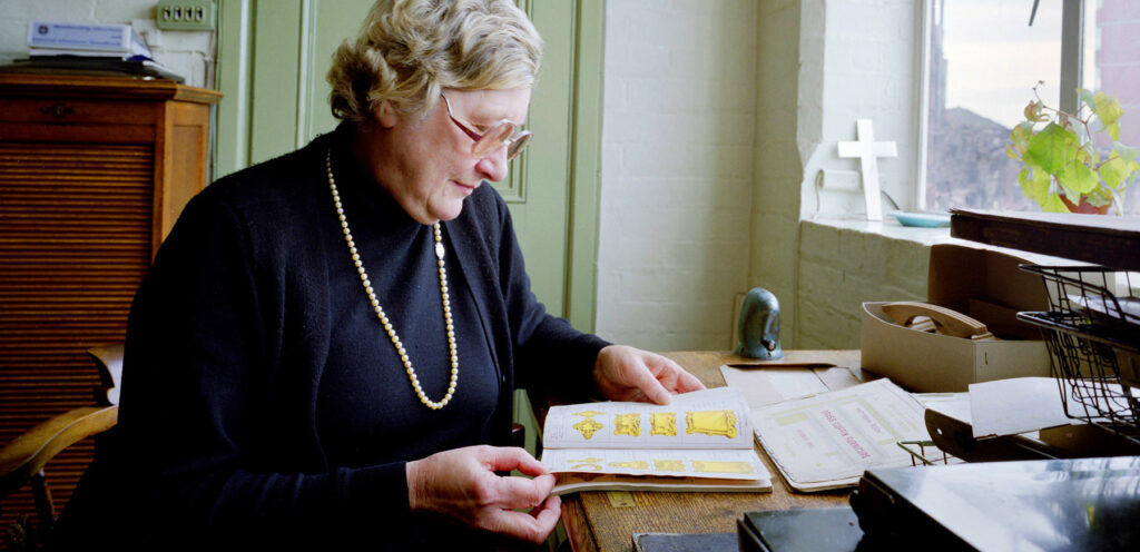 Joyce Green, grey hair wearing a necklace and dark clothing sits at a desk in the office looking through a Newman Brothers' catalogue