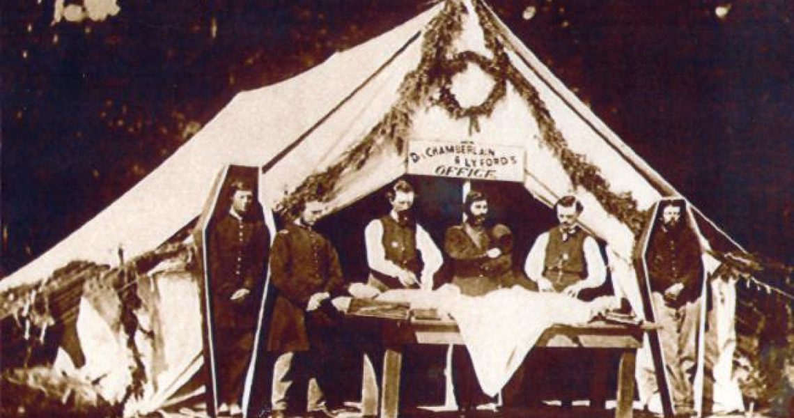 Civil war embalming taking place outside a white tent. There are simple six sided coffins standing on both sides of the tent with soldiers stood inside them.