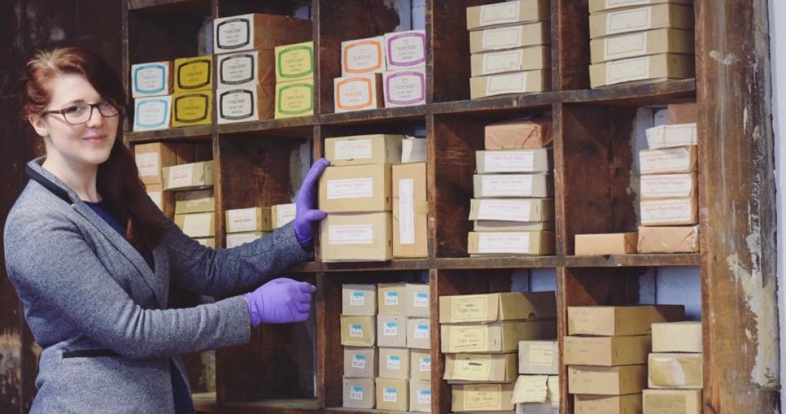 On then left stands a woman with long red hair and grey jacket. Her hand rests on one of the various boxes of coffin handles resting on shelves behind. She wears protective purple gloves.