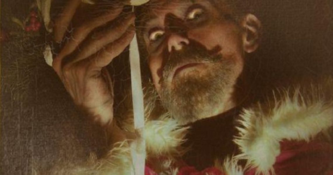 A man with grey moustache and beard wearing Father Christmas clothing looks down at the viewer, he holds a mask in front of his face and is lit from below which gives a spooky feel.